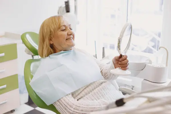 A smiling senior patient checking her teeth in a hand mirror